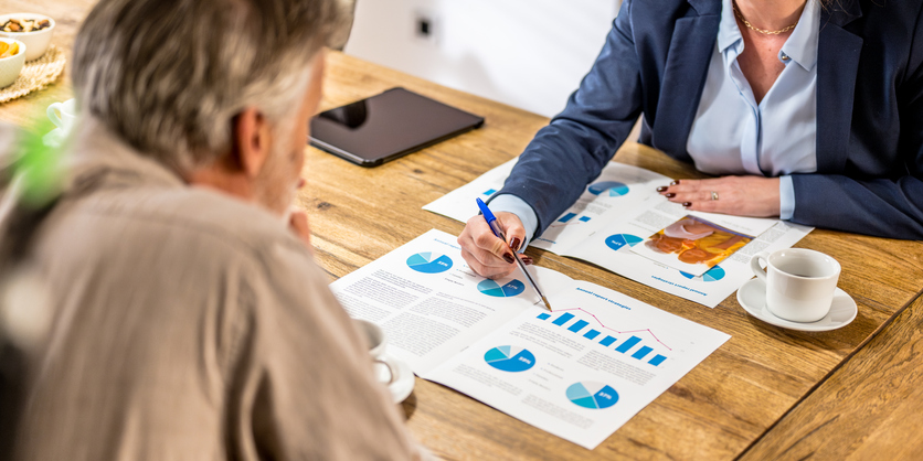 Market research analyst pointing with pen at graphs on documents and explaining senior market research benefits to a client.